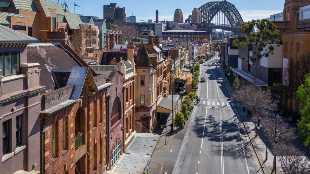 Uma rua repleta de edifícios históricos leva à Sydney Harbour Bridge sob um céu azul claro e com tráfego mínimo.