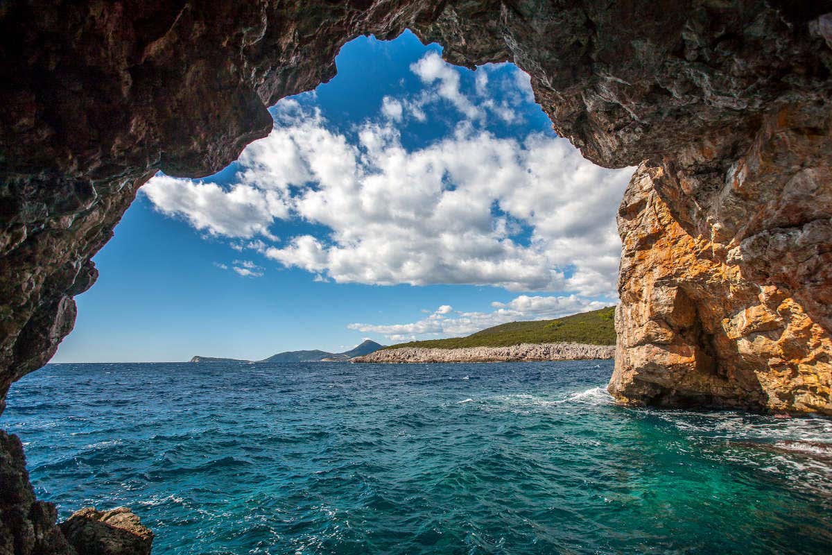 Vista de dentro de uma caverna rochosa, conhecida como Gruta Azul em Perast, com vista para um mar azul com colinas distantes sob um céu cheio de nuvens dispersas, para representar o que fazer em Montenegro.