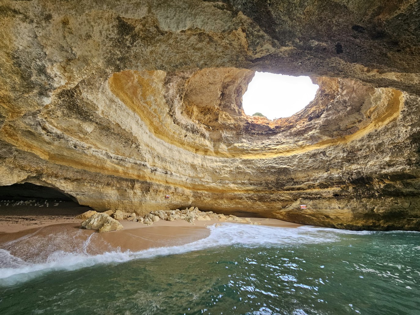 Caverna com uma grande abertura circular acima, praia de areia no interior e ondas chegando à costa, iluminadas por luz natural.