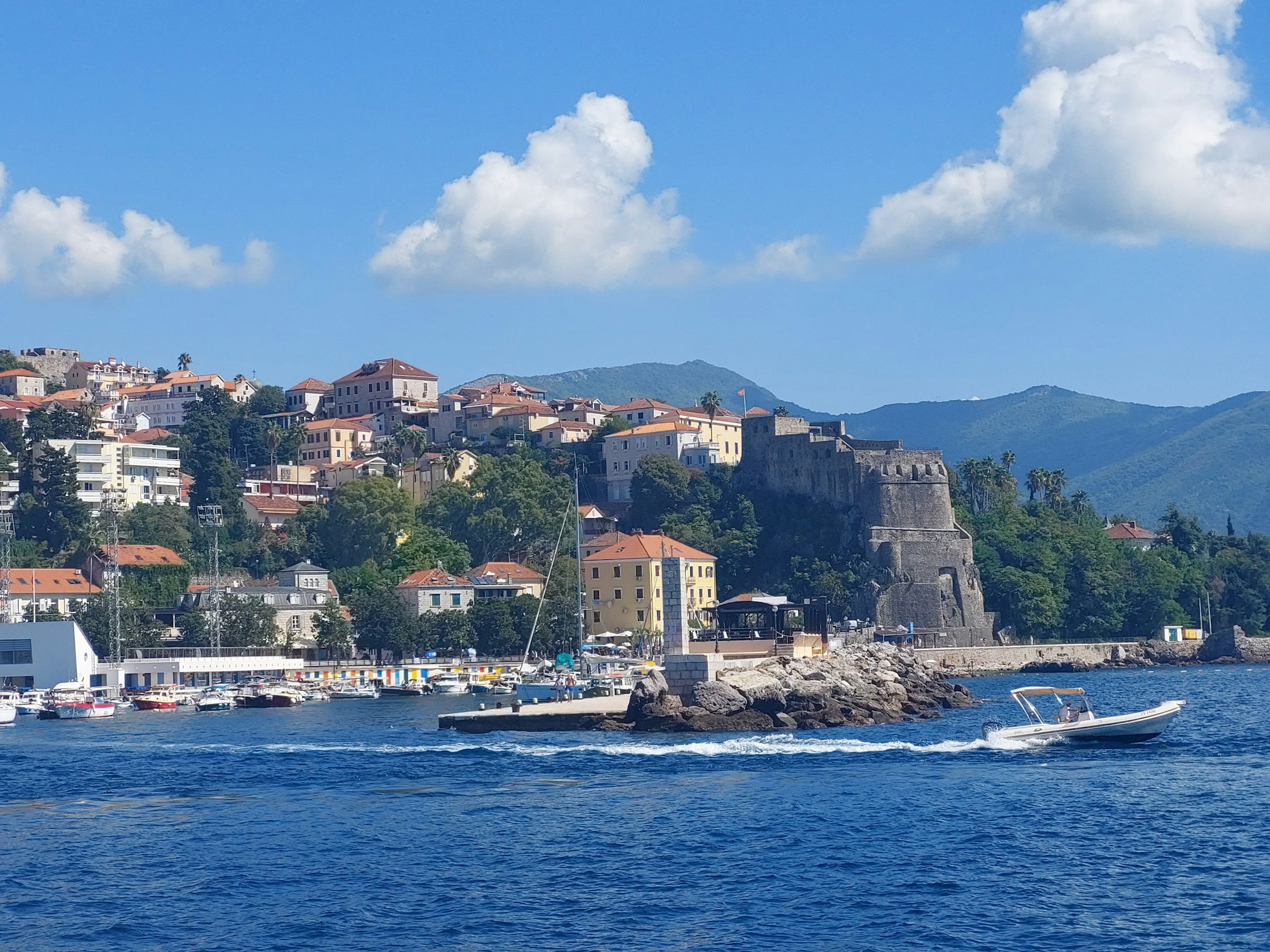 Um pequeno barco navega rapidamente por um mar azul vibrante na cidade costeira de Herceg Novi, com edifícios coloridos e uma fortaleza histórica de pedra, para representar o que fazer em Montenegro.