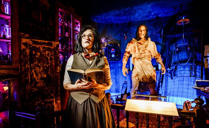 Uma pessoa fantasiada segura um livro em uma sala mal iluminada com uma figura grande e robusta ao fundo.