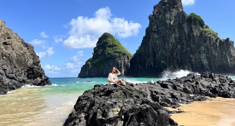 Uma pessoa sentada em rochas negras perto do oceano, com penhascos íngremes e céu azul ao fundo.