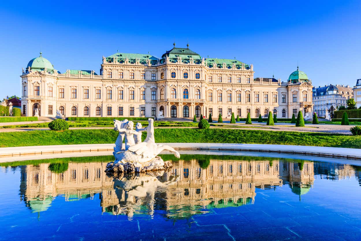 Um palácio barroco histórico com cúpulas verdes e estátuas ornamentadas, refletido em um lago cristalino, sob um céu azul brilhante.
