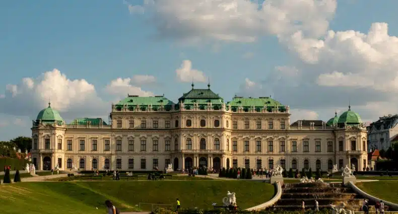 A imagem mostra o Palácio Belvedere em Viena, Áustria, com um jardim bem cuidado e visitantes caminhando pelo caminho. A imagem ilustra o post sobre pontos turísticos de Viena.