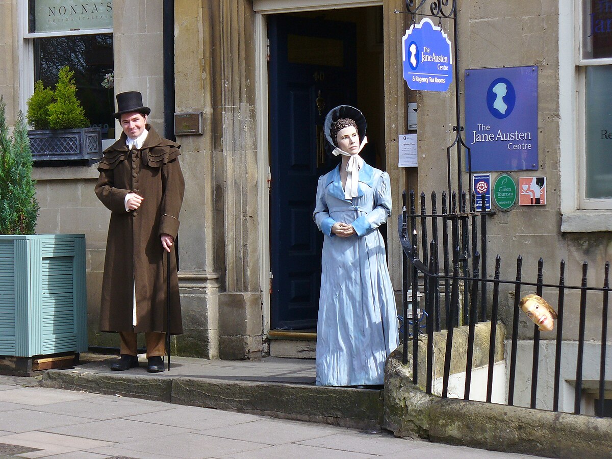 Duas pessoas em trajes de época estão do lado de fora do Jane Austen Centre em Bath. Uma usa um casaco marrom, a outra um vestido azul. Ótima opção de o que fazer em Bath.