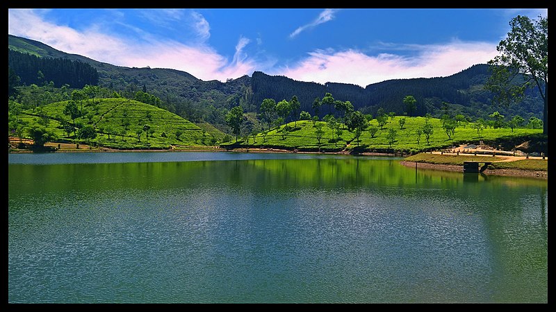 Um lago tranquilo com águas refletivas, cercado por colinas verdes e montanhas distantes sob um céu azul com nuvens finas, em Peradeniya e Matale.