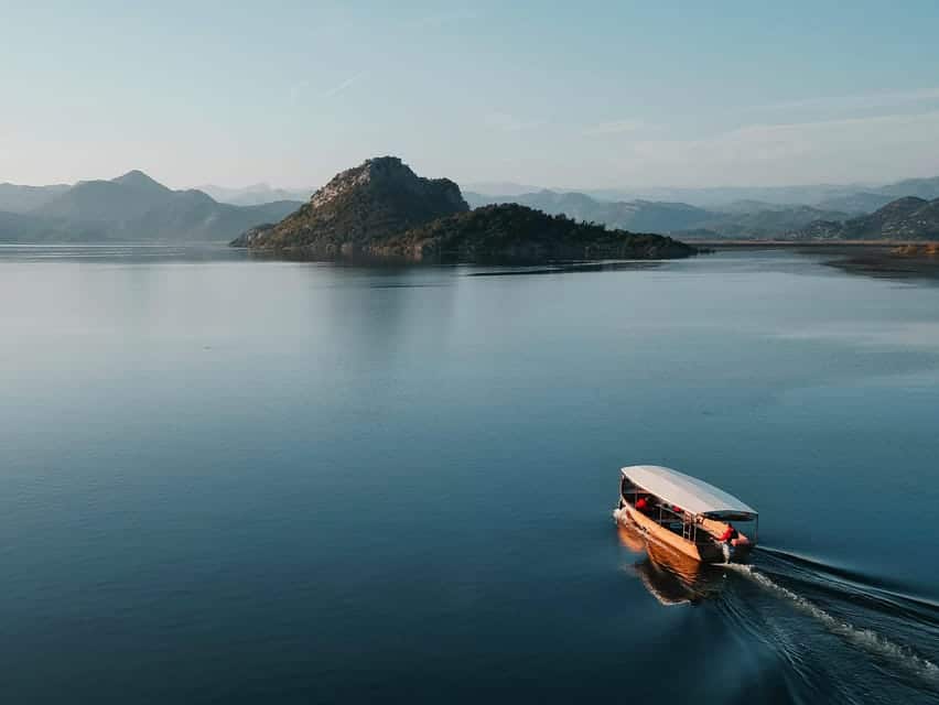 Um barco com cobertura navega em no Lago Skadar, em Montenegro, indo em direção a uma pequena ilha com colinas ao longe, sob um céu limpo.