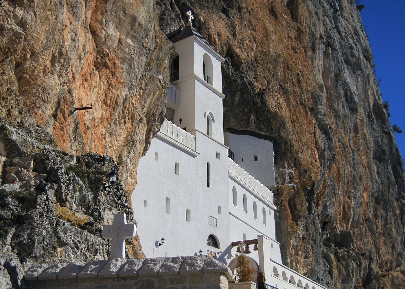 Mosteiro branco aninhado em um penhasco rochoso, trata-se do Monastério de Ostrog, sob um céu azul brilhante, com cruzes visíveis e detalhes arquitetônicos.