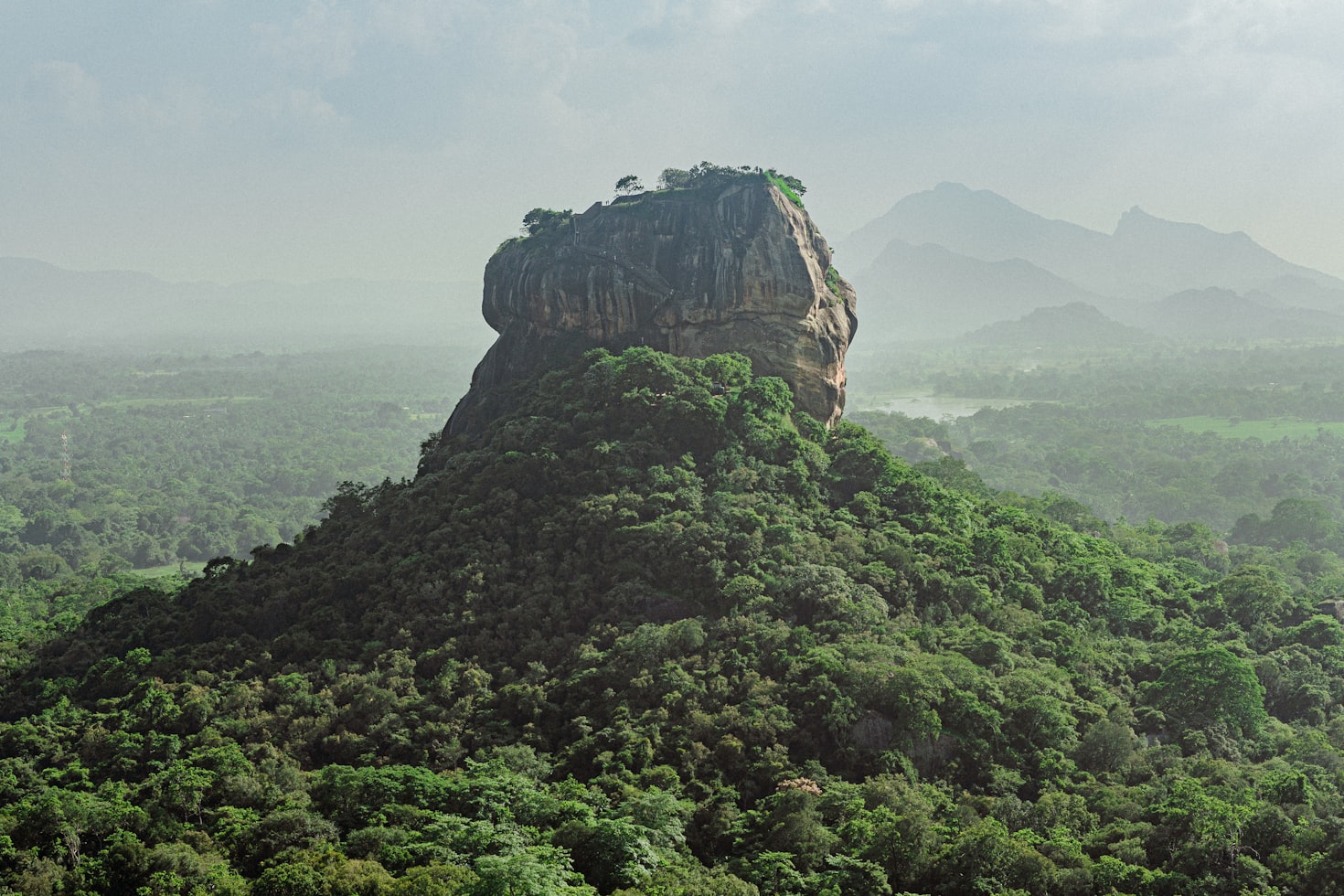 Uma grande formação rochosa com árvores no topo ergue-se em meio a uma densa floresta verde, com montanhas distantes e nebulosas ao fundo, em Sigiriya. Representa o que fazer no Sri Lanka.