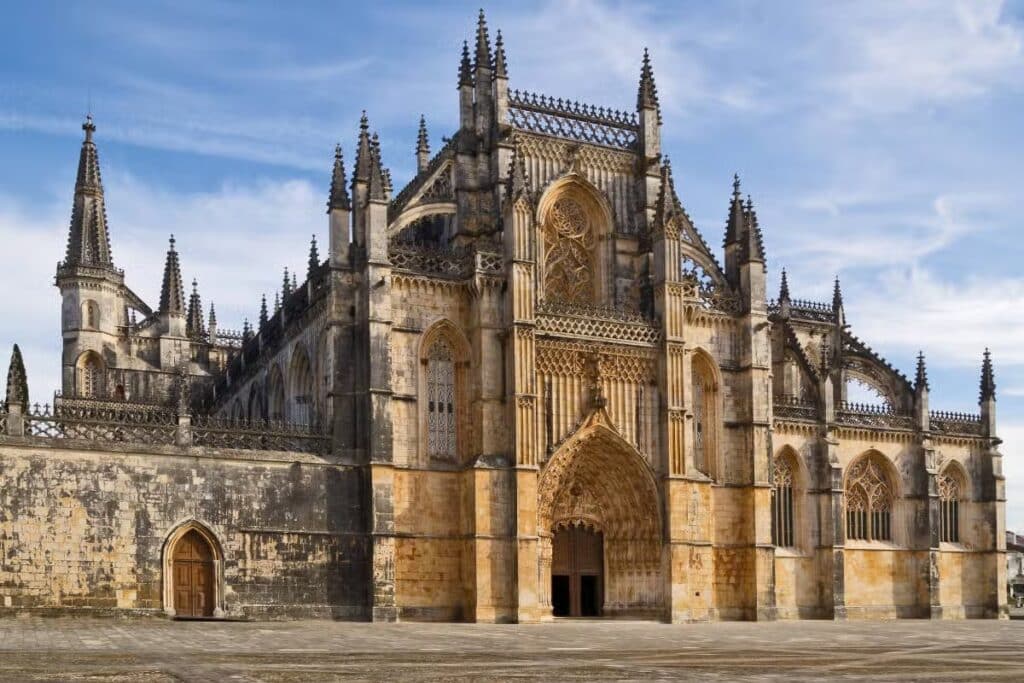 Mosteiro da Batalha em estilo gótico com torres e arcos ornamentados contra um céu azul, com uma grande entrada em arco e paredes de pedra desgastadas.