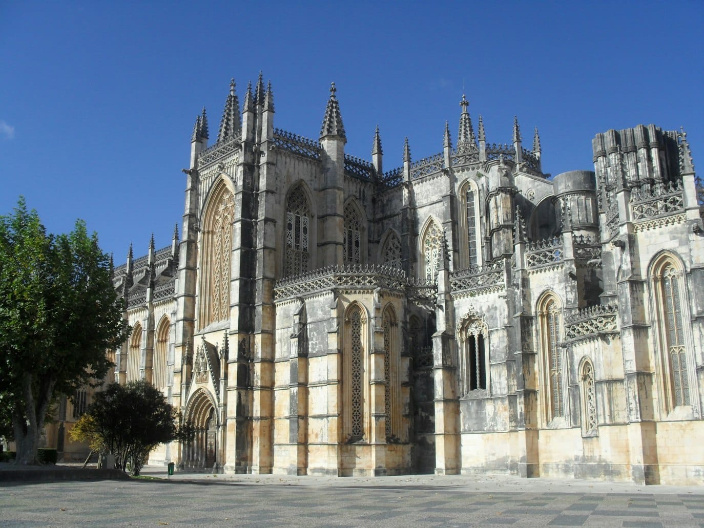 Mosteiro da Batalha em Portugal de pedra em estilo gótico, com torres ornamentadas e janelas em arco, contrastando com um céu azul claro e uma árvore à esquerda.