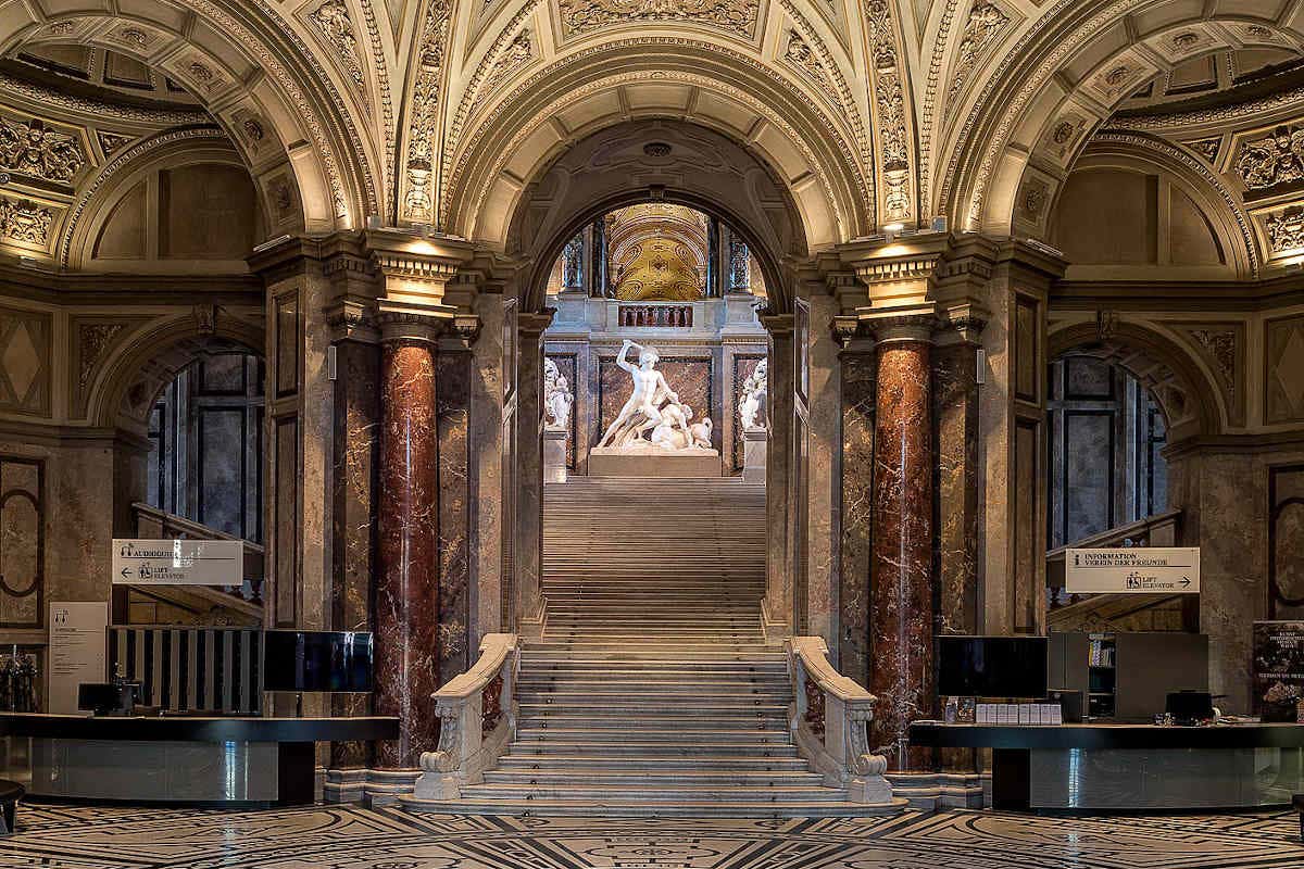 Grande escadaria em um museu com arcos e colunas ornamentados, levando a uma exposição de estátuas no topo, sob tetos intrincados. Esse é o Museu de História da Arte, um dos pontos turísticos de Viena.