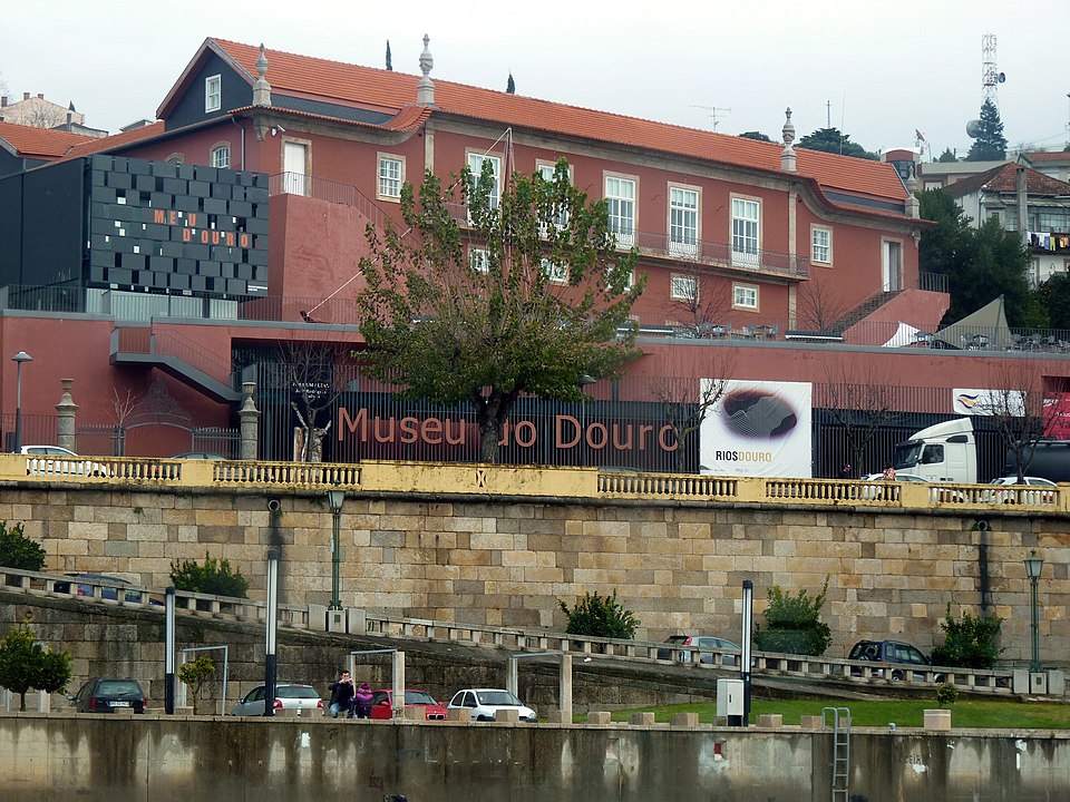 Um edifício de tijolos vermelhos abriga o Museu do Douro. Um outdoor é visível acima dos carros estacionados ao longo de um muro de pedra.
