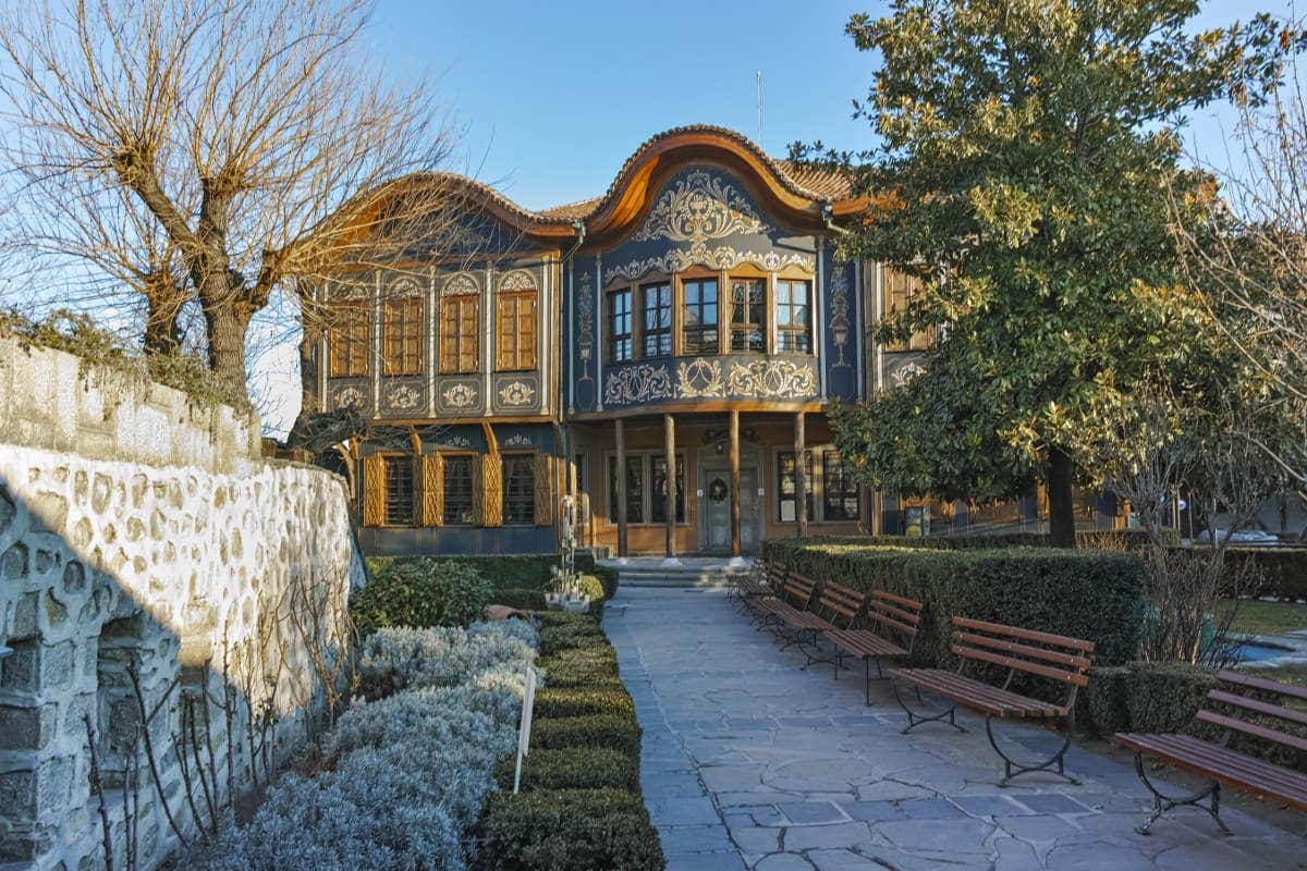 Edifício histórico com fachada ornamentada e bancos de madeira, cercado por jardins bem cuidados, sob um céu azul claro. É o Museu Etnográfico, ilustrando o que fazer em Plovdiv.