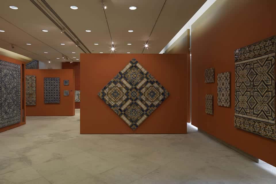 Sala de exposição no Museu Nacional do Azulejo (Lisboa) exibindo vários azulejos com padrões complexos em paredes laranja, com holofotes no teto iluminando o espaço.
