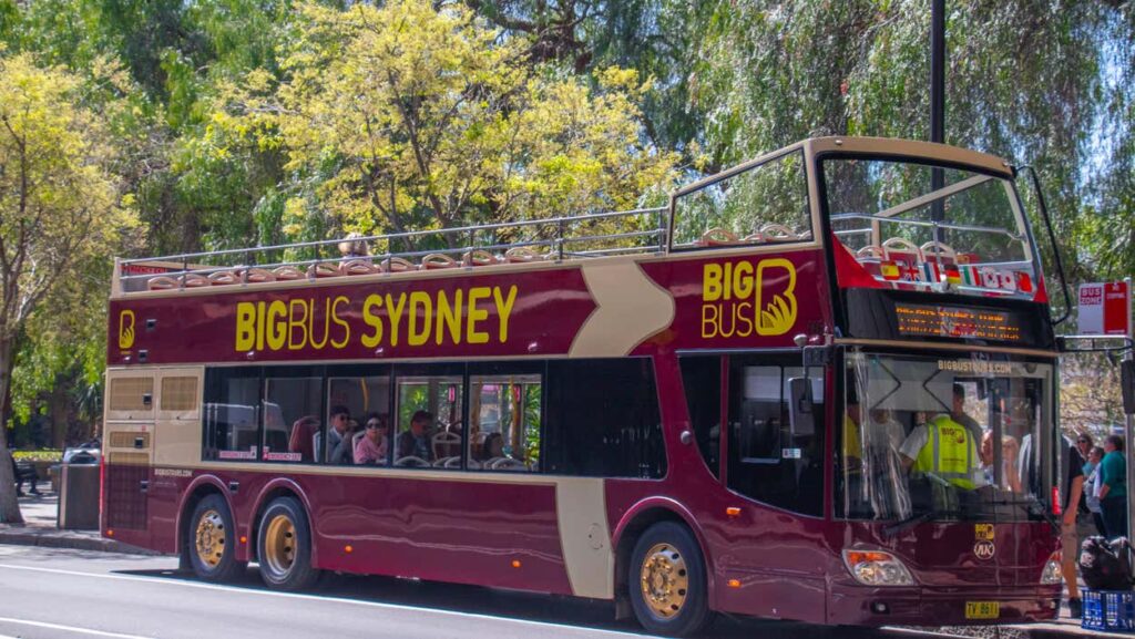 Ônibus de turismo de dois andares denominado "Big Bus Sydney" com passageiros sentados, estacionado em uma rua ensolarada com árvores ao fundo. Imagem para ilustrar post sobre pontos turísticos da Austrália.