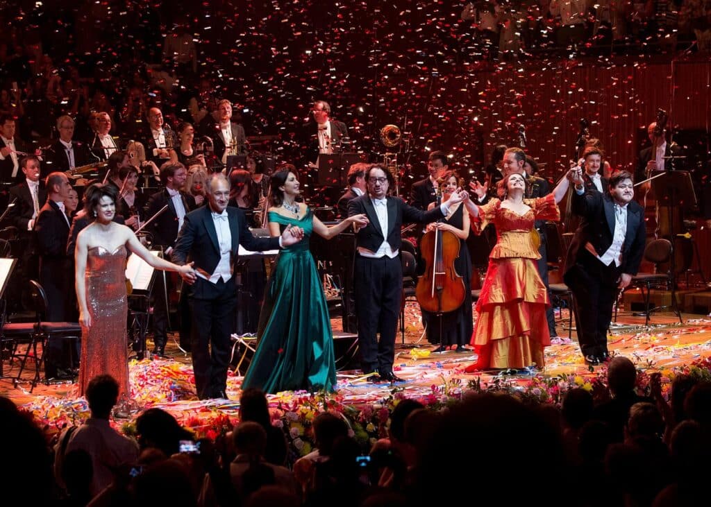 Os artistas no palco da Sydney Opera House dão as mãos durante uma ovação de pé, com uma orquestra sinfônica atrás deles e confetes caindo de cima.