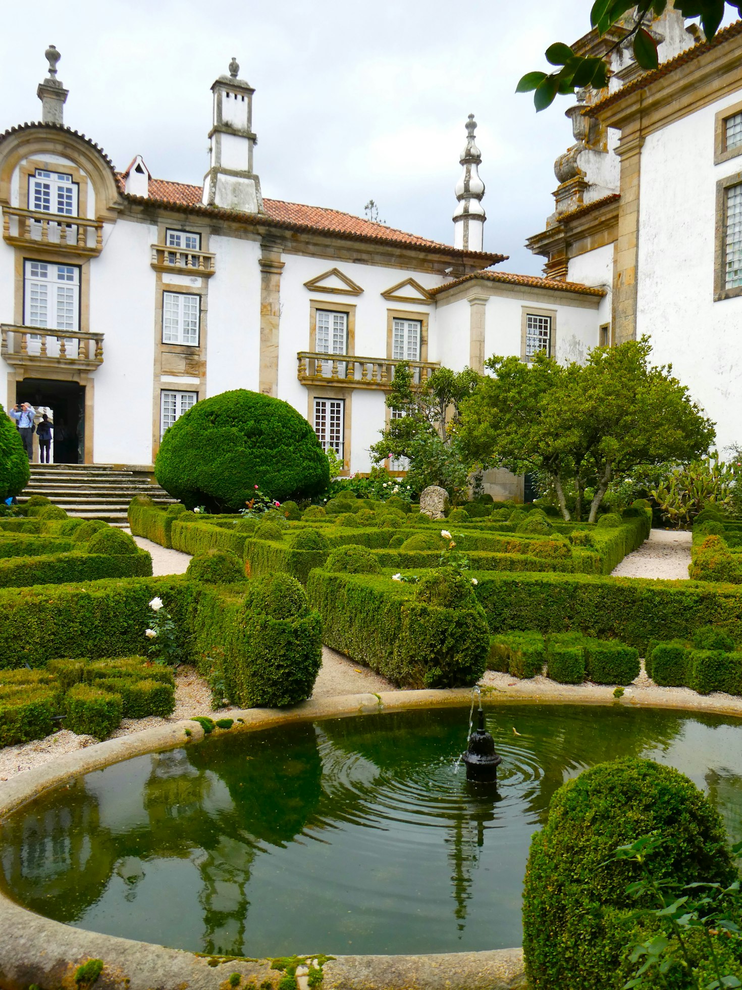 Palácio de Mateus em Portugal, um jardim formal com sebes bem cuidadas, uma pequena fonte circular e um edifício histórico com várias janelas e colunas ao fundo.