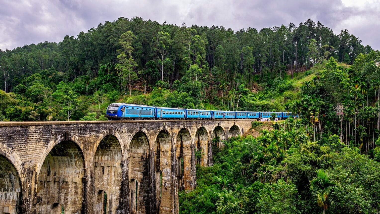 Um trem azul na Ponte dos Nove Arcos cruza um antigo viaduto de pedra cercado por uma floresta verdejante, com um céu nublado. Representa o que fazer no Sri Lanka.
