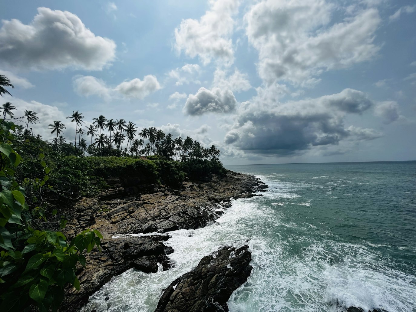 Litoral rochoso de Tangalle com ondas quebrando contra a praia. Palmeiras alinham a borda da terra e nuvens enchem o céu acima. Representa o que fazer no Sri Lanka.
