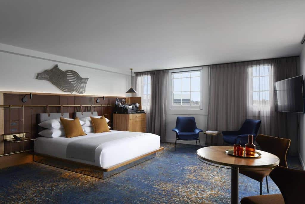 Quarto de hotel com cama king-size, duas cadeiras azul-marinho, mesa redonda e TV na parede. Janelas grandes com cortinas transparentes. Foto do Pier One Sydney Harbour.