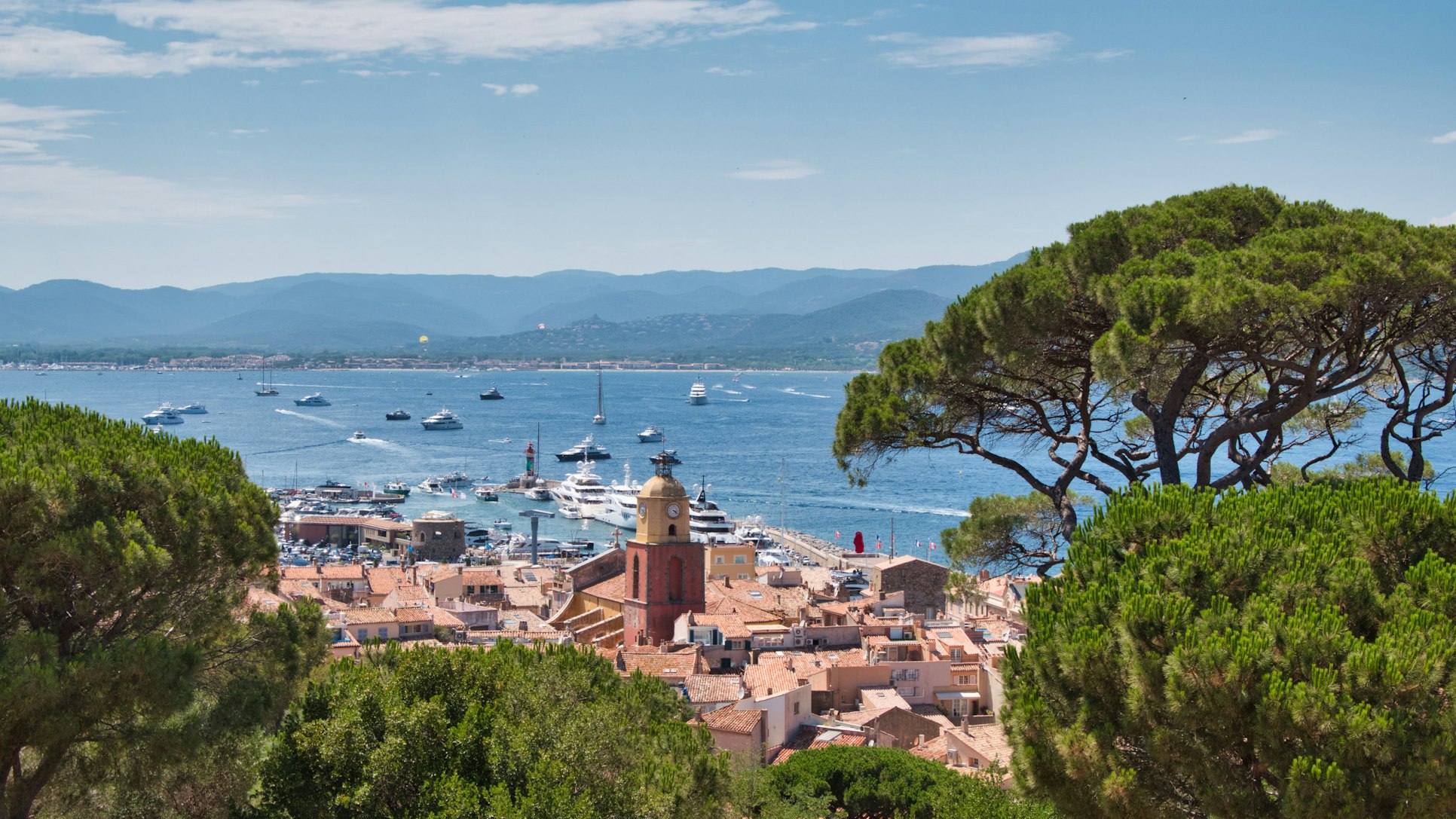Vista de uma cidade costeira com telhados de terracota, uma torre de igreja, barcos no porto e montanhas ao fundo em um dia claro. Essa é Saint-Tropez, uma das dicas de o que fazer no sul da França.