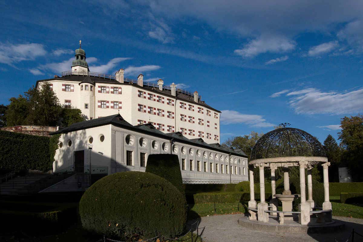 Um castelo branco com inúmeras janelas, uma torre em forma de cúpula e um jardim com arbustos bem cuidados e um mirante de pedra sob um céu azul claro.