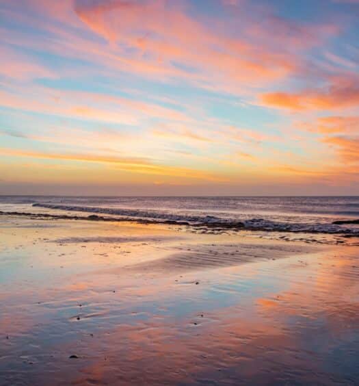 Um casal caminha pela praia de Jericoacoara ao pôr do sol, com nuvens coloridas refletindo na areia molhada e ondas calmas ao fundo. A imagem ilustra o post sobre as melhores praias do Nordeste.