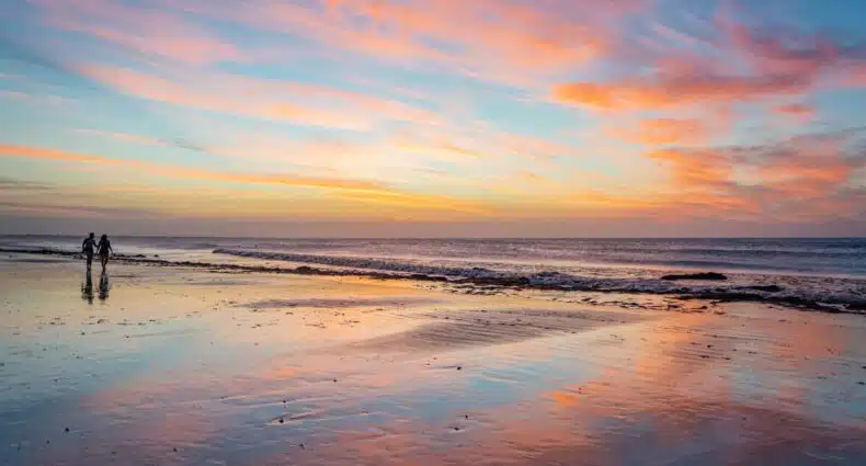 Um casal caminha pela praia de Jericoacoara ao pôr do sol, com nuvens coloridas refletindo na areia molhada e ondas calmas ao fundo. A imagem ilustra o post sobre as melhores praias do Nordeste.