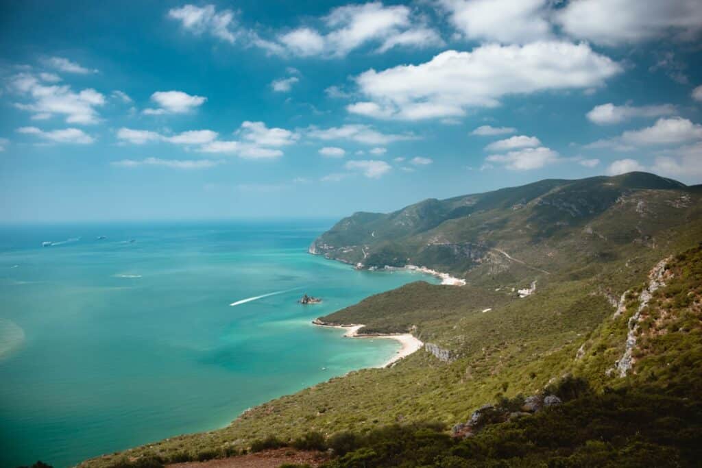 Vista aérea de uma paisagem costeira com mar azul-turquesa, praias de areia, colinas verdes e algumas nuvens dispersas no céu.
