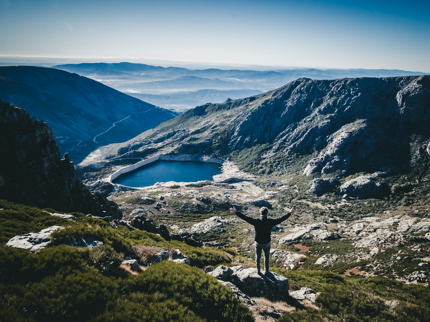 Pessoa em pé sobre uma rocha, braços estendidos, observando uma paisagem montanhosa com um lago e colinas distantes sob um céu azul na Serra da Estrela em Portugal
