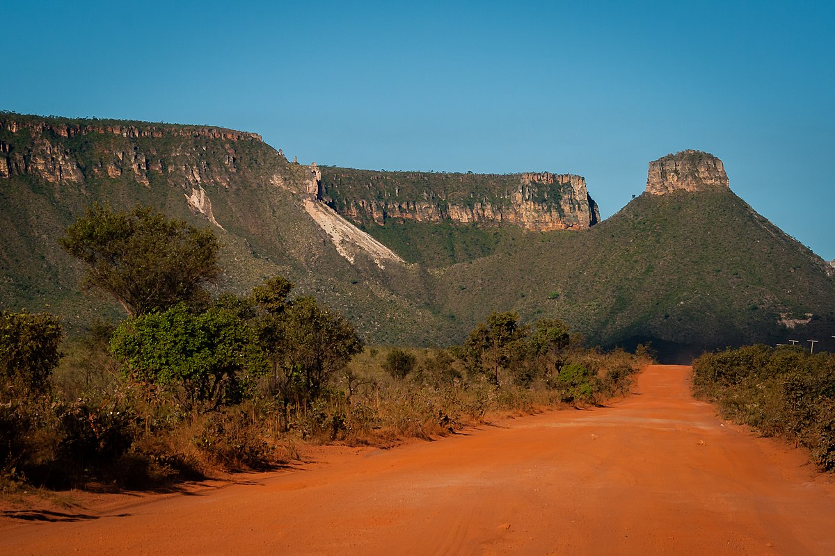 Uma estrada de terra vermelha leva a uma cadeia de montanhas de topo plano sob um céu azul claro, cercada por vegetação esparsa e arbustos.