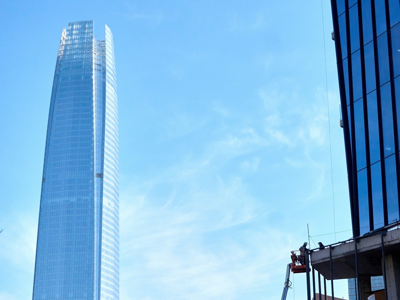 Arranha-céu alto e moderno do Sky Costanera com fachada de vidro à esquerda e outro edifício em construção à direita, contra um céu azul.