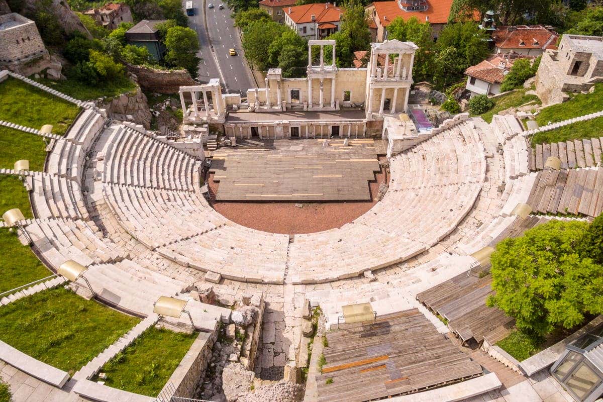 Vista aérea de um antigo anfiteatro romano com assentos de pedra e palco, cercado por vegetação e estradas próximas com carros. Ilustra o que fazer em Plovdiv.