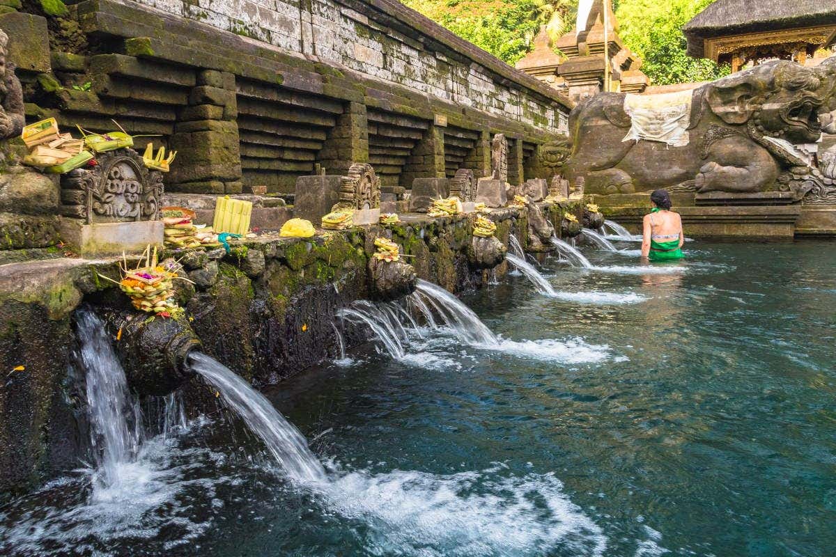 Uma pessoa em uma piscina com fontes de pedra, cercada por oferendas e esculturas, perto de um antigo muro de pedra. É o templo Tirta Empul. Está ilustrando o post sobre o que fazer em Ubud.