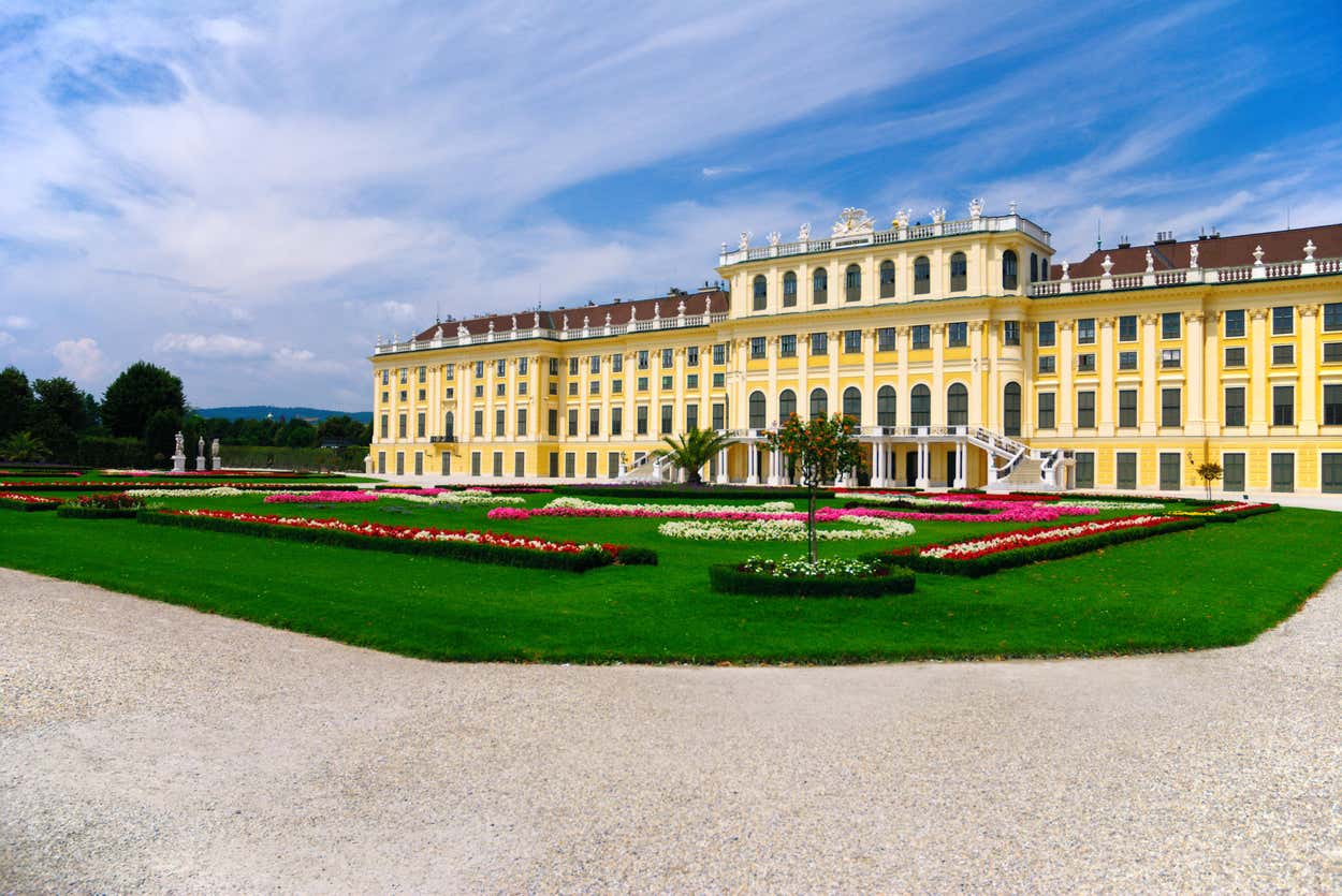 Um grande palácio amarelo claro com janelas brancas, decorações ornamentadas no telhado e jardins bem cuidados sob um céu azul com nuvens.