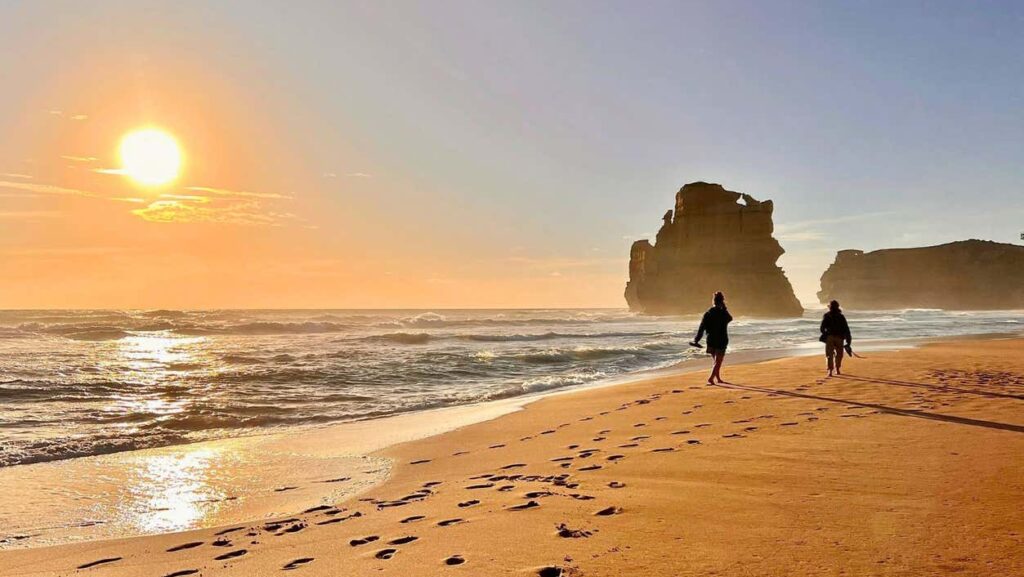 Duas pessoas caminham ao longo da praia Twelve Apostles com pegadas, ondas do mar e uma formação rochosa ao pôr do sol.