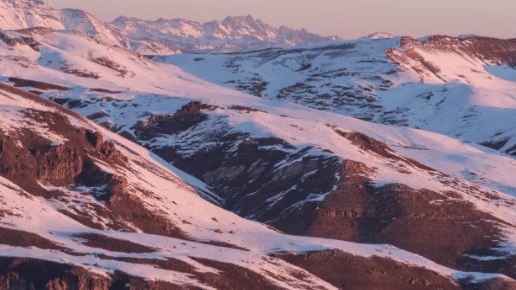 Paisagem montanhosa coberta de neve ao amanhecer com tons rosa e roxo no céu. Terreno rochoso parcialmente visível, no Vale do Nevado.