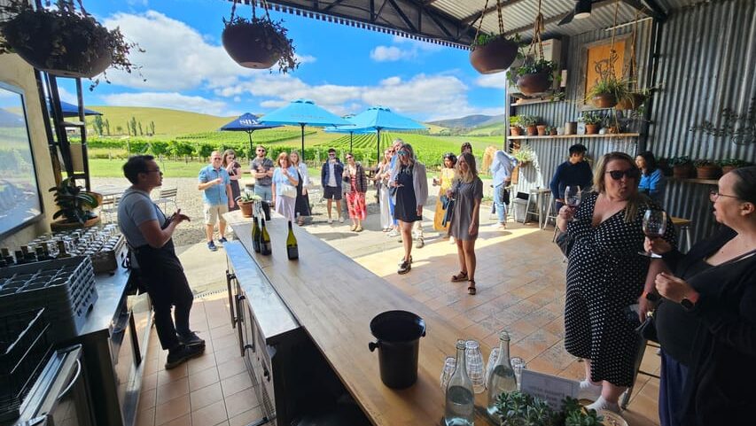 Pessoas em um evento de degustação de vinhos em um ambiente rústico ao ar livre com vinhedos ao fundo, sob um céu parcialmente nublado em Yarra Valley. Imagem para ilustrar post sobre pontos turísticos da Austrália.