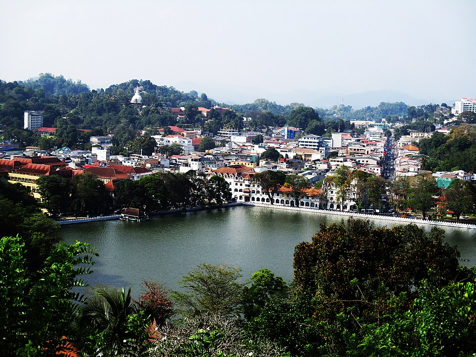 Vista aérea de uma cidade de Kandy com um grande lago em primeiro plano, cercada por edifícios, árvores e colinas sob um céu limpo. Representa o que fazer no Sri Lanka.