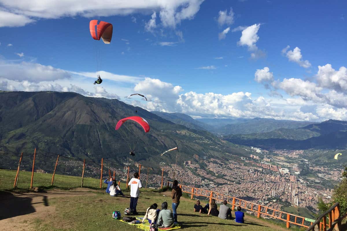 Pessoas sentadas em uma colina gramada observando parapentes voando sobre uma paisagem urbana e montanhas sob um céu azul com nuvens.