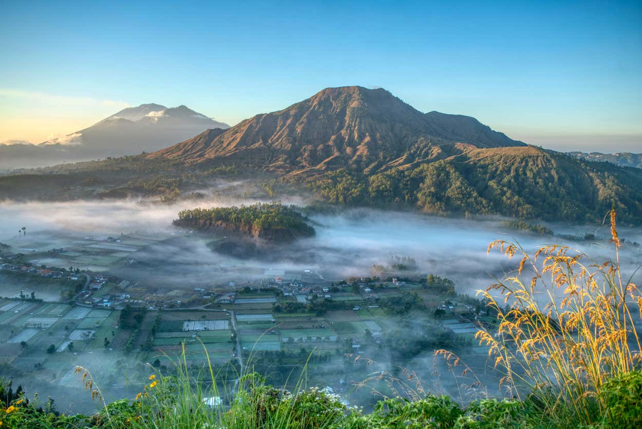 Uma vista panorâmica do Monte Batur com campos e vilas cobertos de neblina em primeiro plano, contra um céu azul claro. Está ilustrando o post sobre o que fazer em Ubud.