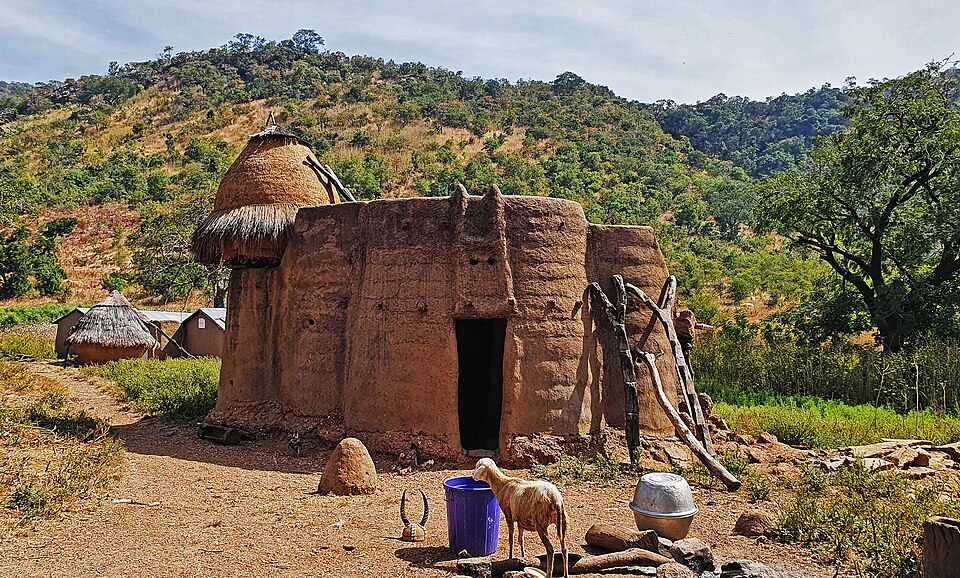 Casa redonda de barro com telhado de palha, escada de madeira, cabra, baldes e potes; colinas e árvores ao fundo. Que pode ser visitado com o eSIM Togo.