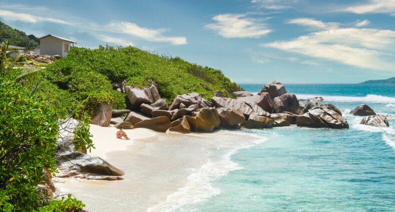 Uma pessoa sentada em uma praia arenosa com pedras, arbustos verdes e ondas azuis do oceano sob um céu parcialmente nublado em La Digue, Seychelles. Que pode ser visitado com o eSIM Seychelles.