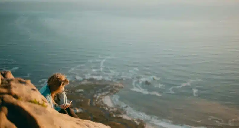 Pessoa sentada em um penhasco rochoso em Lion's Head Peak, Cidade do Cabo, com vista para o mar, usando um smartphone. O litoral e as ondas do mar são visíveis abaixo. Para ilustrar o post de eSIM África do Sul.