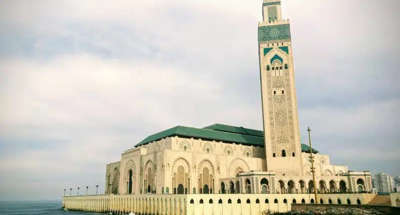 A grande Mesquita de Hassan II com um alto minarete fica ao lado do oceano, com ondas batendo em sua base sob um céu nublado. Que pode ser visitada com o eSIM Casablanca.