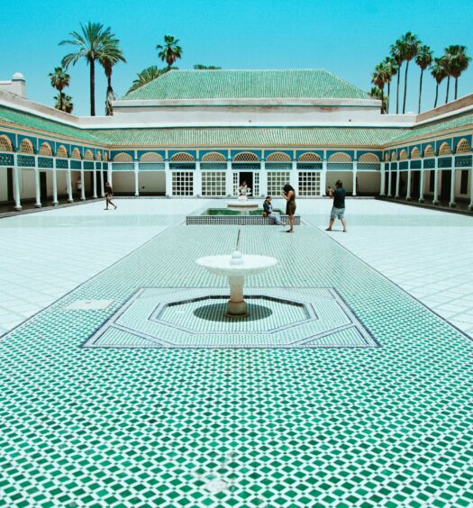 Um grande pátio de azulejos com uma fonte central, passarelas em arco e palmeiras ao fundo sob um céu claro em Palácio da Bahia, que pode ser visitado com o eSIM Marrakesh.