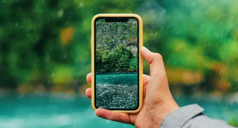 Uma pessoa segura um smartphone capturando uma paisagem de floresta e rio. A tela do celular exibe a mesma imagem de um ângulo diferente do Puerto Varas, Los Lagos, Chile, que pode ser visitado com o eSIM América do Sul.