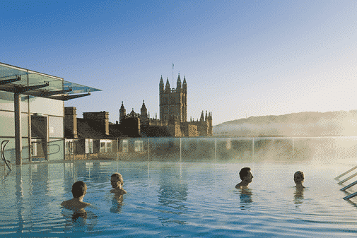 Quatro pessoas em uma piscina ao ar livre com vapor subindo no Thermae Bath Spa. A Abadia de Bath com pináculos é visível ao fundo sob um céu limpo.