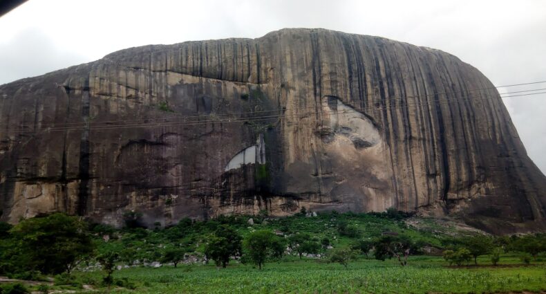 Uma grande colina de Zuma Rock rochosa com estrias verticais, vegetação verde na base e um céu nublado acima. Pode ser visitada com o eSIM Nigéria.